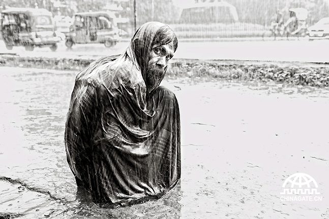 达卡雨季，一个流浪汉在倾盆大雨中瑟缩在人行道上。意料之外的大雨让这个城市流浪汉们的生活更加困难重重。