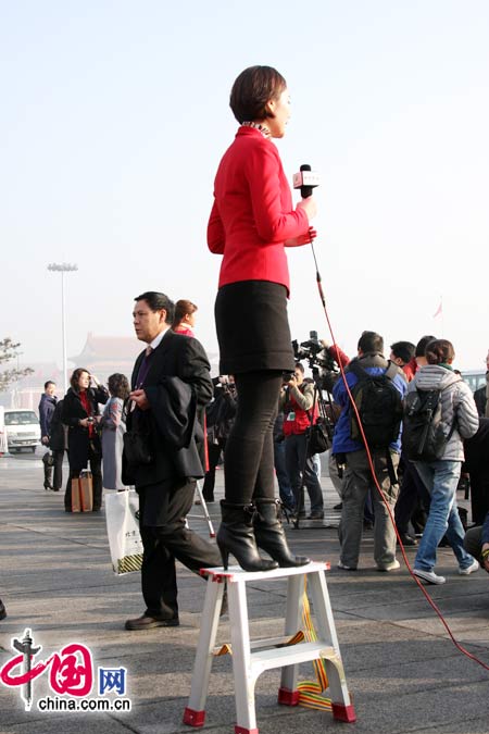 A TV reporter is reporting what she has seen from standing high on a simple stool, and furthermore, she wears high heel shoes. 