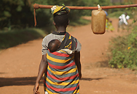 A young mother is carrying her child on the back and a hand hoe toggled with a bucket from farm back home.