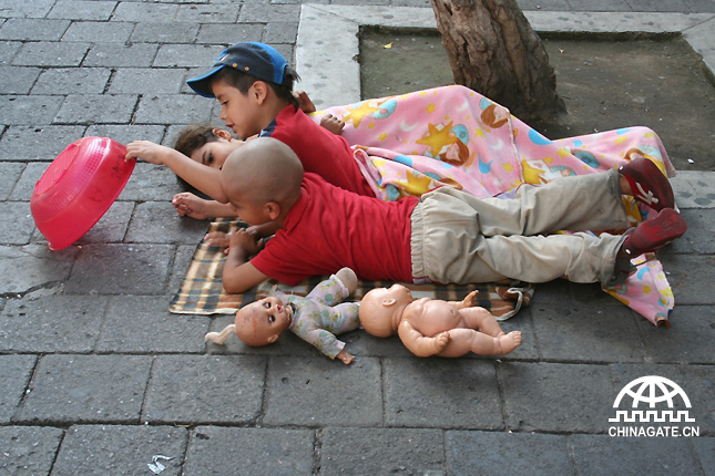 Three kids are playing on the floor on the street. People seem to adapt to poverty, and society gets used to it, unless they see better quality life ways to live.